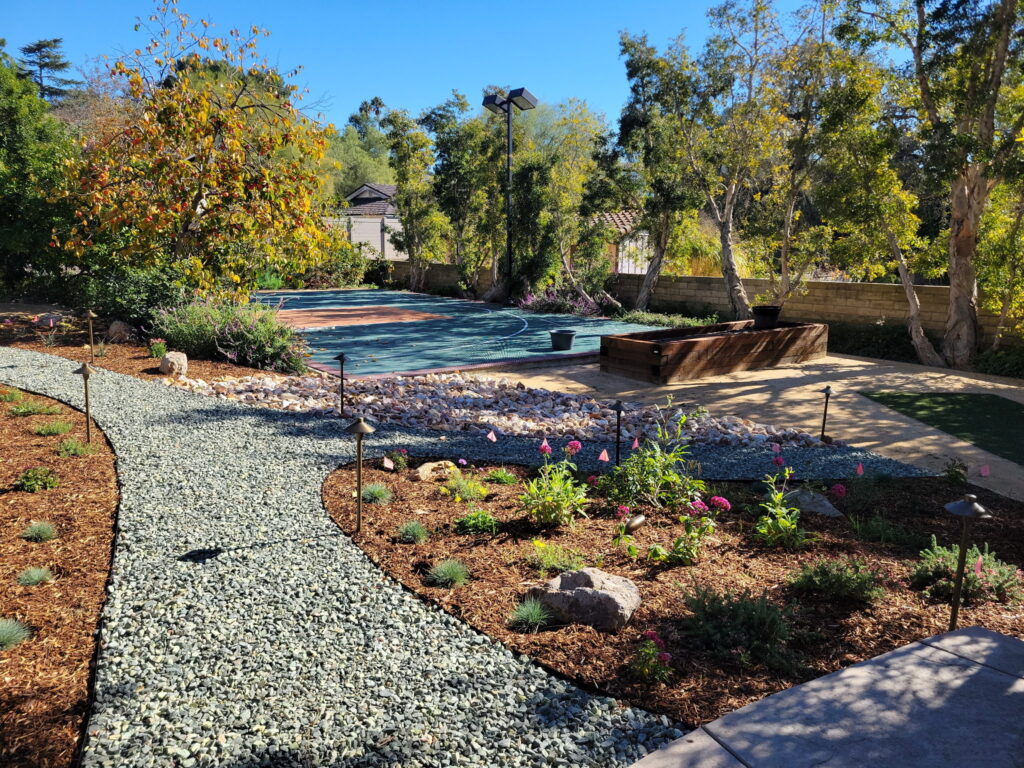 Curved gravel path with small lights winds through a landscaped garden of shrubs, trees, and a wooden planter box under a clear blue sky.