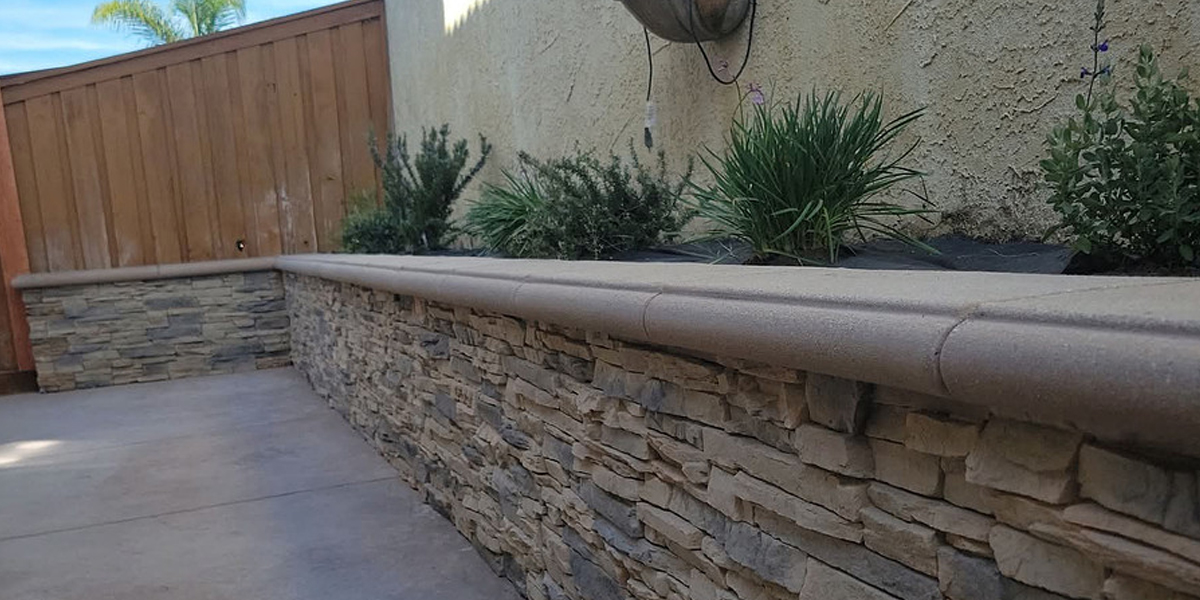 Stone veneer retaining wall with a rounded concrete cap, planter along a concrete patio.