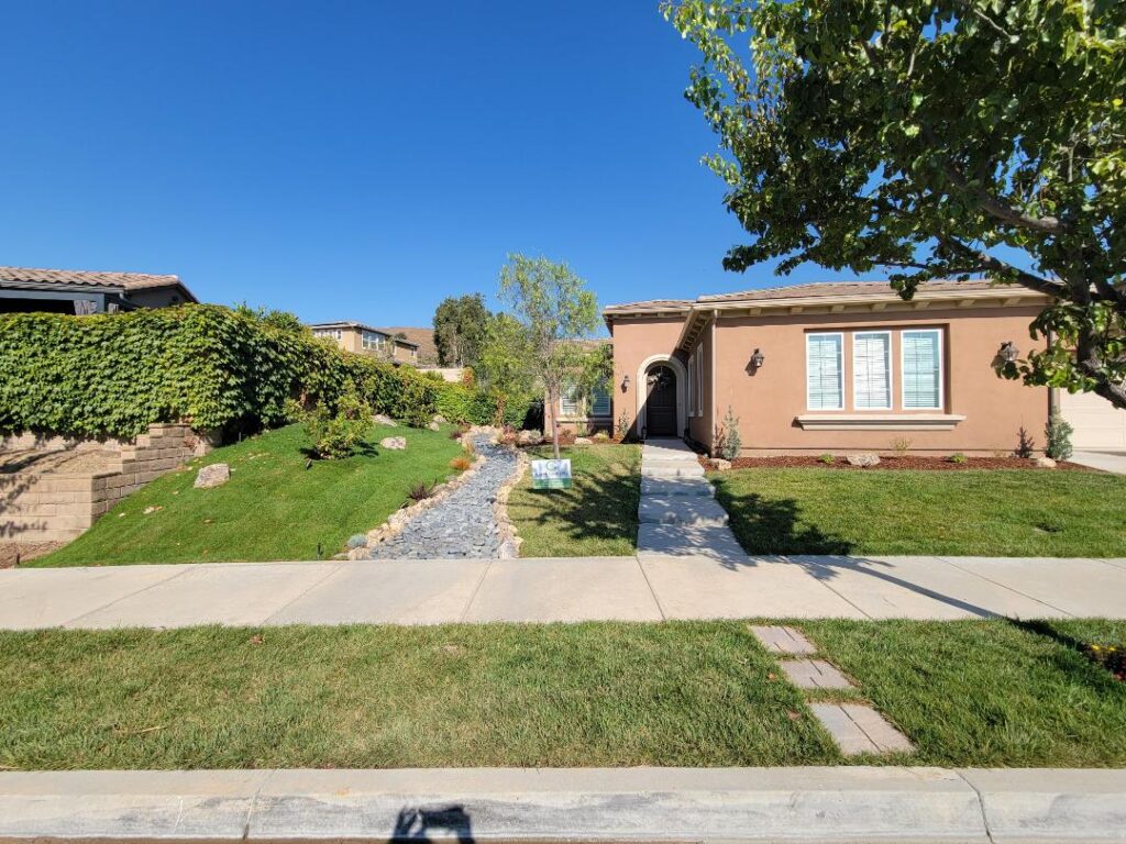 Front view of a single-story tan house with an arched entry, green lawn, and a stone pathway leading to the front door under a clear blue sky (for sale sign nearby).