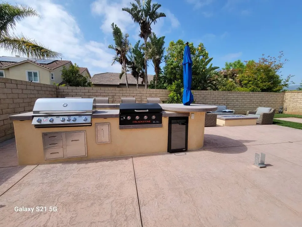 Outdoor kitchen: built-in grill, Blackstone griddle, mini fridge, and counter space in a sunny backyard with a brick wall and palm trees.