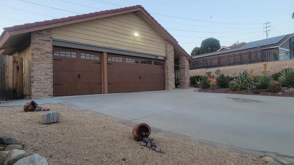 Three-car garage home with stone pillars, beige siding, and a concrete driveway; desert landscaping with rocks and cacti in the yard.