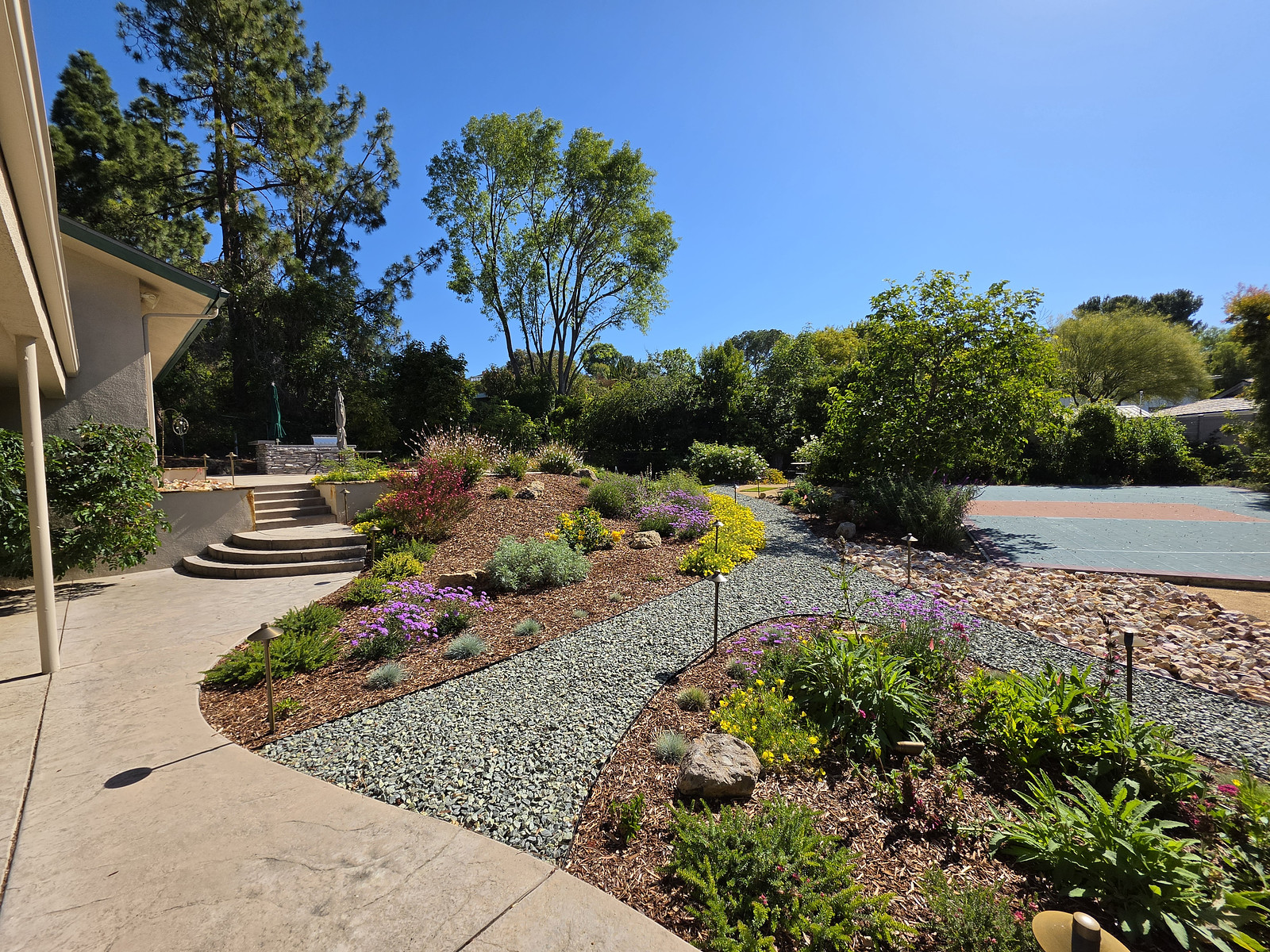 Sunny backyard with stone steps, raised garden beds, and colorful flowering plants along gravel paths by the house edge.