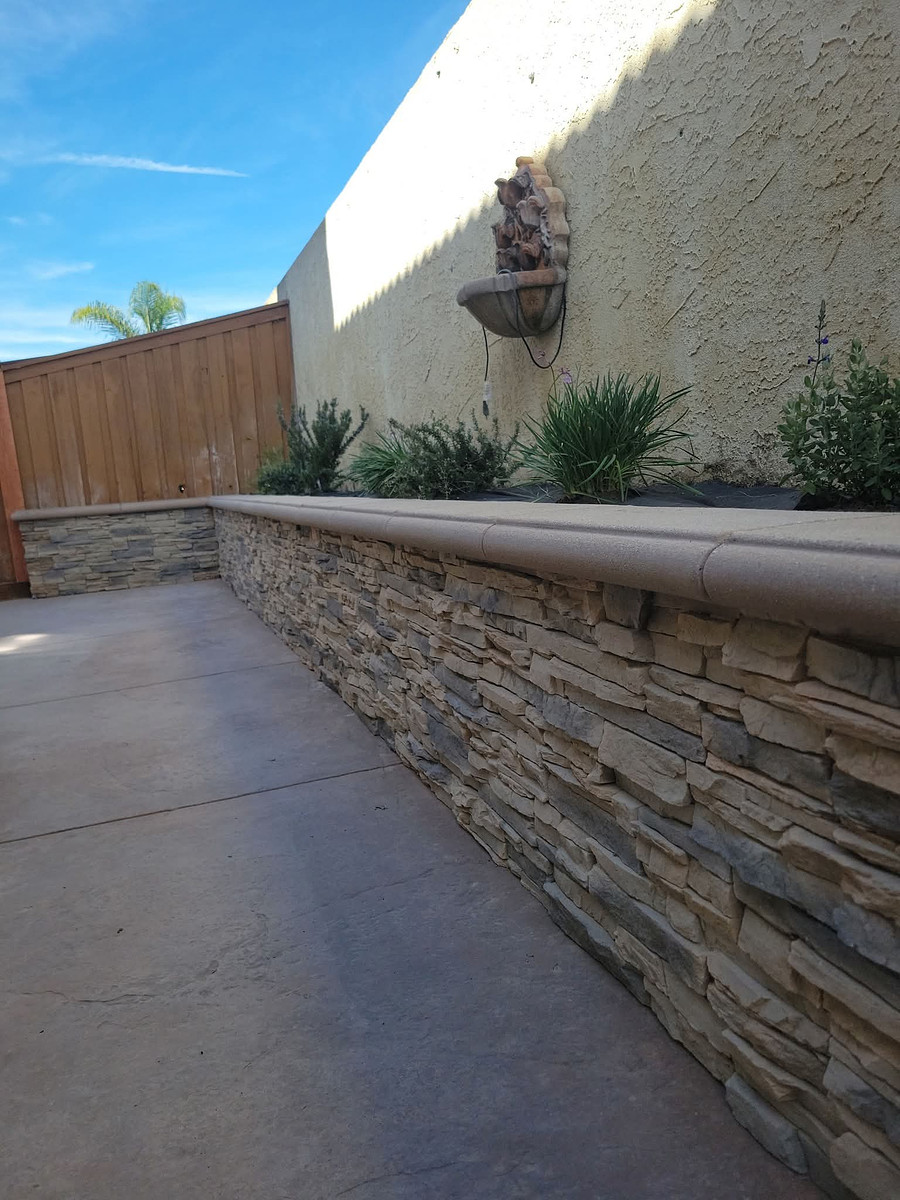 Concrete walkway beside a stone-faced retaining wall with a planter and a wall-mounted fountain on a beige stucco wall, blue sky above.