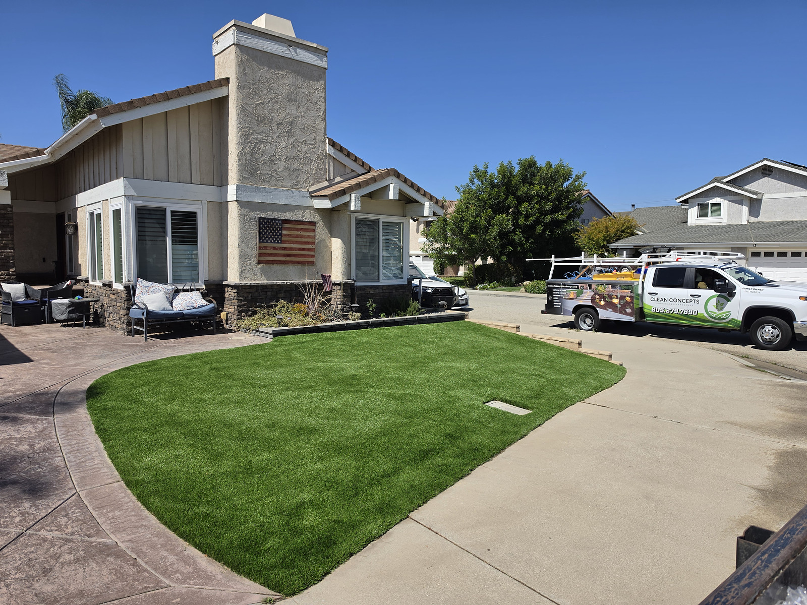 Front yard of a suburban house with green lawn and a patio seating area; a service van is parked in the driveway.
