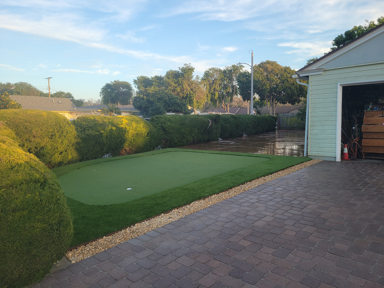 Backyard putting green beside a brick-paved driveway, with neatly trimmed hedges and an open shed showing storage inside.