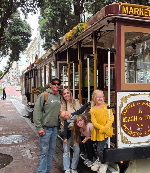 Family of five posing with a vintage trolley car decorated with flowers on a city street.